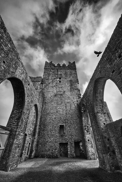 A black and white, low-angle shot of the ruins of Tintern Abbey, showing stone walls reaching towards a cloudy sky with a bird flying overhead.