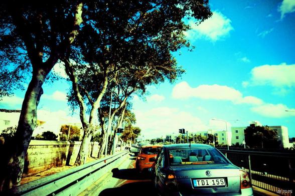 Farbfoto mit übersättigten und etwas ins Exotisch-Nostalgische gedrehten Farben. In Bildmitte sind Autos auf einer Straße in Malta unterwegs. Links Leitplanen and dahinter schüttere Bäume. Rechts ein Metallgeländert und weiter entfernt Häuser. Rechts oben knallblauer Himmel mit weißlichen Wolken.