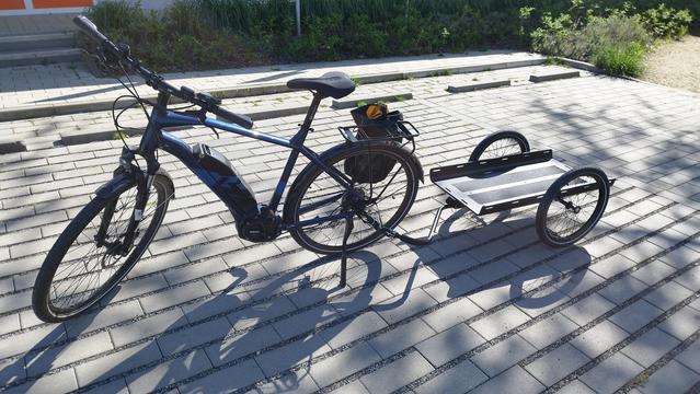 Ebike mit einem Lastenanhänger. Er ist eine gerade Ladefläche die vorne und hinten offen ist. Seitlich eine Kante die paar cm hoch ist. Hat Schlitze für Spanngurte.
