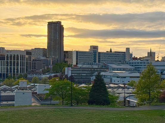 This image captures a cityscape at sunset. The sky is painted with hues of orange and yellow, casting a warm glow over the scene. Dominating the center is a tall, cylindrical building, its silhouette stark against the bright sky. Surrounding it are various modern structures, including a prominent glass-fronted building and others with distinctive architectural designs. In the foreground, a grassy area with trees and a pathway leads the eye toward the city. The overall mood is serene and reflective, with the setting sun adding a tranquil ambiance to the urban landscape.