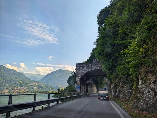 The image captures a scenic view of a road winding alongside a lake, framed by lush greenery and mountains in the distance. An old stone archway tunnel is visible, adding a historical element to the landscape. A vehicle is seen driving on the road, emphasizing the scale of the natural surroundings. The sky is partly cloudy, casting soft shadows and highlights across the scene.