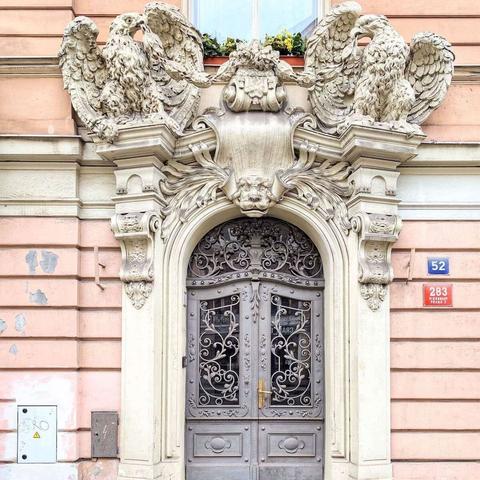 A street-level view of an ornate Art Nouveau doorway in Prague featuring gray metal double gates with intricate swirling patterns. The arched entrance is framed by a light-colored stone archway adorned with elaborate carvings, including two large sculpted eagles perched above, flanking a central cartouche with a lion's head. To the right of the entrance, a blue plaque displays the number "52," and below it, a red plaque reads "283" The building's facade is a muted pink with horizontal lines.