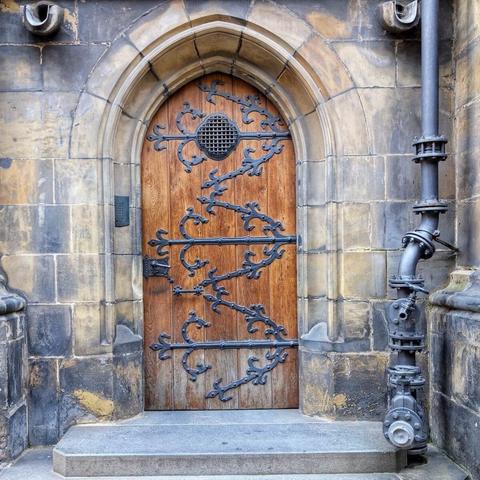 A weathered wooden arched door with intricate black ironwork, set in a stone archway. The door features a small, round grated window at the top and decorative iron hinges and straps with stylized floral or vine patterns. The stone of the archway shows signs of age and weathering. A dark metal pipe runs vertically along the right side of the stone frame. A stone step leads up to the doorway.