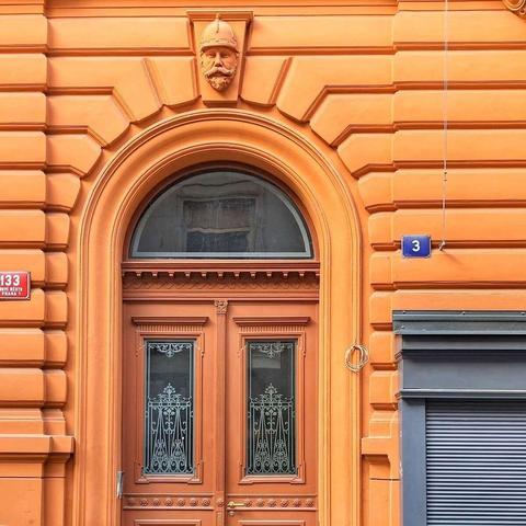 A street-level view of a warm orange building facade featuring a wooden arched double door with decorative metalwork in the glass panels. The doorway is framed by a stylized arch with horizontal grooves, and a carved stone head wearing a helmet is positioned above the arch. To the left of the entrance, a red plaque reads "133 NOVÉ MĚSTO PRAHA 1." To the right, a blue plaque displays the number "3." A portion of a gray roll-down shutter is visible on the right side of the frame.