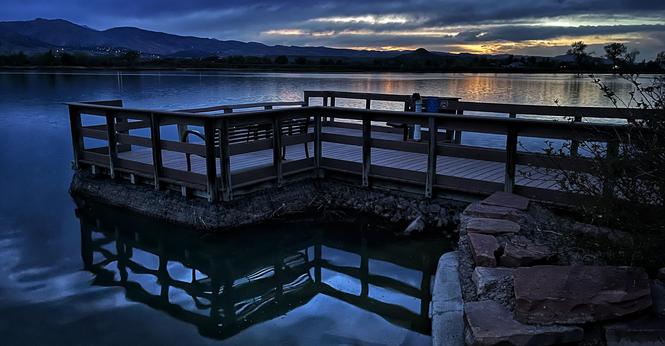 A small wooden pier with sitting benches extending out into a small lake, its wavy reflection in the waters below, with mountains and a few house lights on the far side of the lake, and a mostly cloudy sunset sky with a few peeks of golden orange color among the clouds.