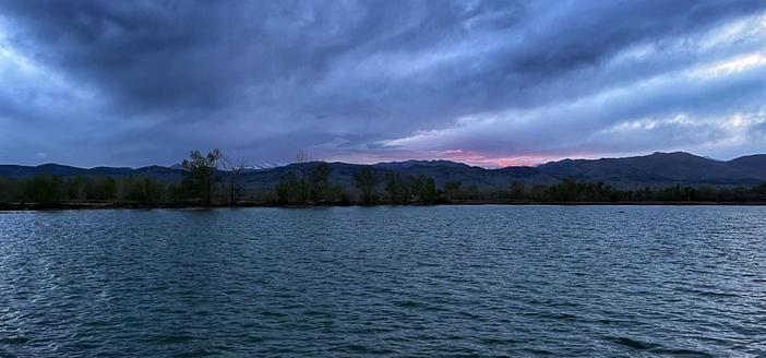 View across a small lake filled with gentle ripples on the surface of the water, trees on the far side and a line of mountains in the far distance (with some last season snow visible in spots), with mostly cloudy skies and a few hints of pinkish sunset color in the clouds.
