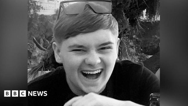 A black and white picture of the Harvey Willgoose, who is laughing. He wears sunglasses on the top of his head, trees and plants can be seen in the background.