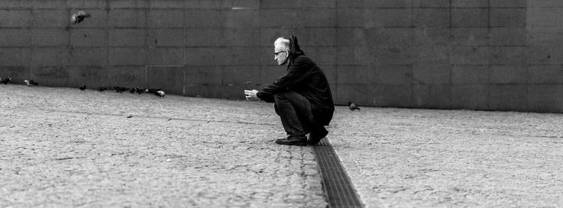 The image depicts a person in a black and white setting, crouching on a cobblestone ground near a wall. The person is wearing a jacket and appears to be holding something. Pigeons are also present on the ground nearby. The composition conveys a sense of solitude or contemplation.