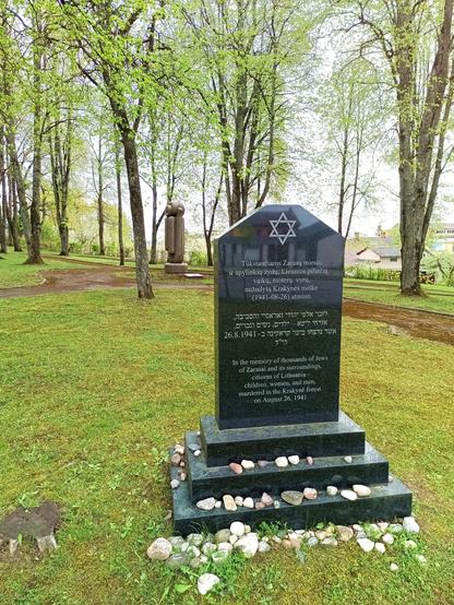 A black memorial stone with a six pointed star at the top stands in a park. A modern art sculpture is further behind. The text on the memorial stone reads: "In the memory of thousands of Jews of Zarasai and its surroundings, citizens of Lithuania - children, women, and men, murdered in the Krakynė forest on August 26, 1941".

The paths are mossy and someone put stones on the steps of the memorial.