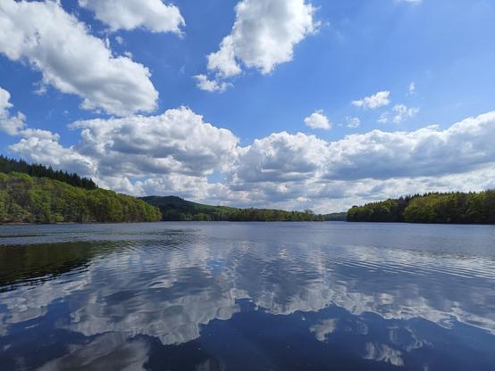 Photographie numérique couleur en format paysage.
Vue d'un lac depuis le bord avec le ciel lumineux et des nuages blancs qui se reflètent dans l'eau très calme.
De part et d'autres, des collines plantées de feuillus près du lac et de sapins sur le sommet.