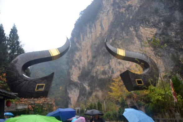 a photo of a gateway,  the gateway is two oxen hons, large tall mountains rising up around it, at the bottom are peoples umbrellas, visitors going to see it
