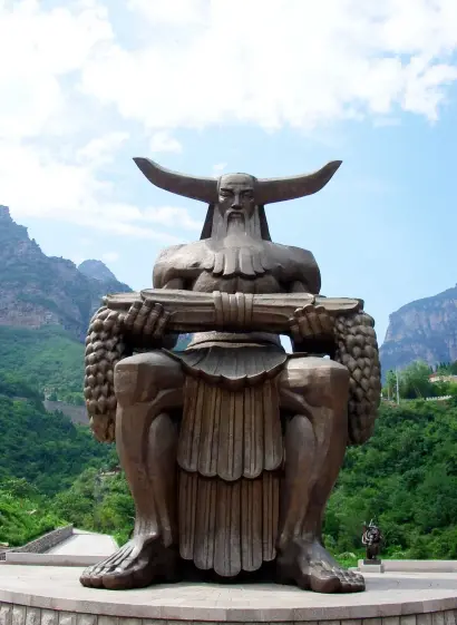 a statue of shennong, seated, he is holding wheat in his hands, a cloudy blue sky and rolling tall mountains behind him