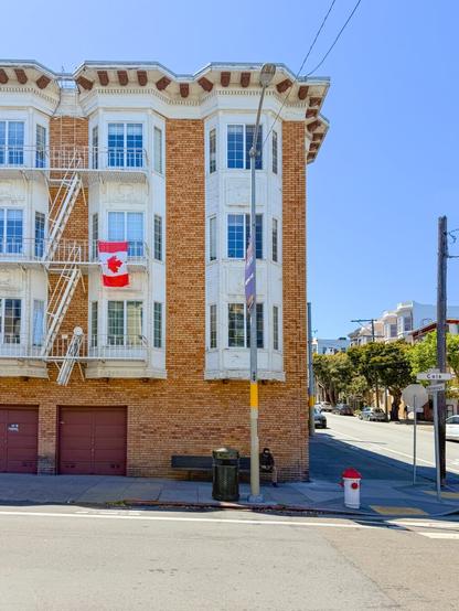 Photograph of a large, Edwardian-era apartment building at the corner intersection of two urban streets. A tan-colored brick veneer covers the building’s façade, interrupted by three columns white-painted bay windows and a zig-zagging, white fire escape ladder. An unfurled, red-and-white Canadian maple leaf flag hangs from the middle fire escape landing. The flag’s colors are echoed in a white-and-red fire hydrant that stands at the curb below. A row of smaller Victorian buildings and a brilliant blue sky are visible in the background.