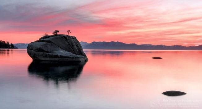 Bonsai Rock in Lake Tahoe, Nevada, is a large granite boulder just off the eastern shoreline. It's name comes from the two small "bonsai" trees growing in cracks at the top of the rock. The photo shows the rock in a mirror still lake with a red-orange sunset and distant mountain peaks in the background. There are two smaller rocks to the right that barely break the surface of the lake.