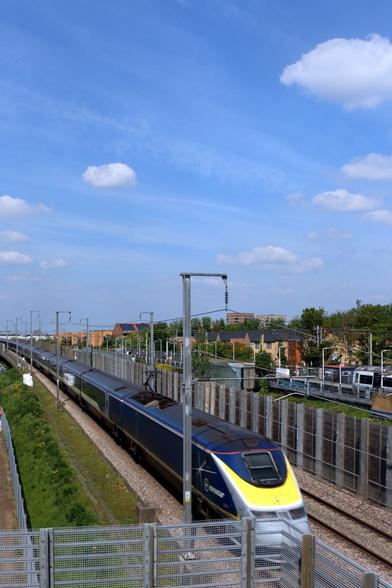 Eurostar train on HS1 captured from footbridge over the line, the yellow face of the driver's can is lofer right while the rest of the train in dark blue livery recedes into the distance centre left. Above is a a blue sky with small white clouds.