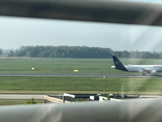 A Lufthansa airplane is seen on a runway with green fields and trees in the background. The image appears to be taken through a window.