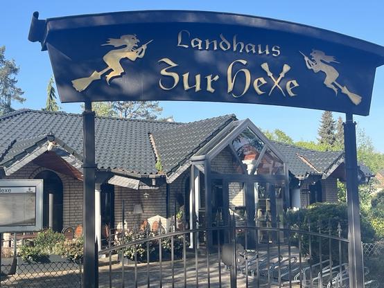 A sign reading "Landhaus zur Hexe" is displayed above a quaint building with a thatched roof. The structure features a mixture of glass and brick, with outdoor seating visible. The surrounding area has greenery and trees, creating a charming atmosphere.