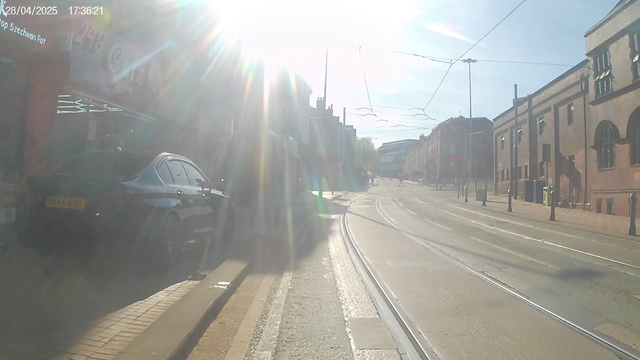 A low quality photograph of a 2 lane street with tram tracks and wide pedestrian pavements on both sides.
A driver has parked a black car on the pavement.