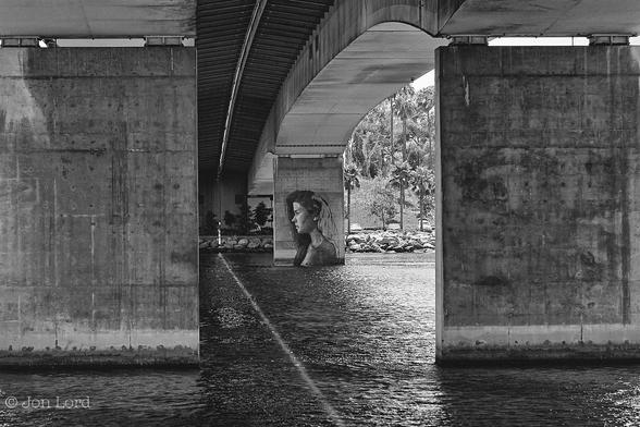 This Is A Black And White Street Photo In Landscape Format. Covering Much Of The Image Is A River Filling A Little Less Than The Lower Half Of The Photo. In The Foreground Are Two Large And Grubby Rectangular Concrete Pillars Supporting An Arched Bridge That Spans The River. On The Far Side Is A Further Pillar On The Right Side Of The Bridge. This Rectangular Pillar Has A Large Piece Of Street Art Painted On Its Surface. This Shows A Young Woman In Profile, From The Shoulders Upwards And Looking To the Left. She Has Long Dark Hair And A Slightly Pointed Nose. There Is A Suggestion That The Art Work Is Not Yet Completed As Some Of The Hair At The Back Of The Head Is In Outline Only. In The Background The Shore Is Made Up With Large Rocks, Behind A Few Parked Cars With Rising Terrain And Palm Trees.
The Location Is On The Banks Of The Los Angeles River In Long Beach, California. The Structure Is The Queensway Bridge (2016).