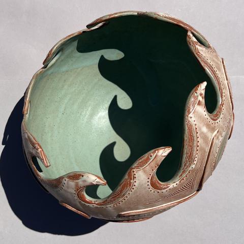 A decorative ceramic bowl featuring a textured, wave-like design along the rim. The interior is a smooth, muted green, contrasting with the intricately detailed outer surface. The photo is square and the background is plain, light gray. Bright, direct sunlight casts a shadow of waves against the inside of the bowl.
