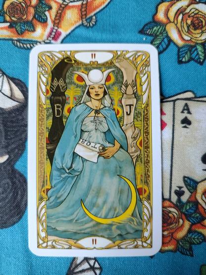 Card from the Golden Art Nouveau Tarot deck. 
The High Priestess-II.
A woman dressed in light blue, with a white crown, holding a scroll. She is sitting between two columns.