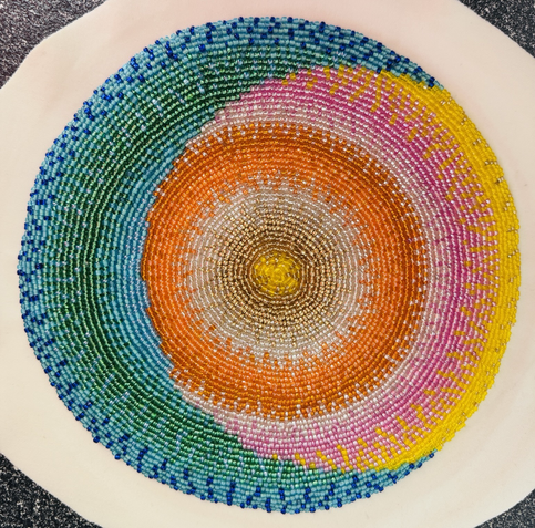 A beaded circle showing a blue crescent shape eclipsing a circle with rows of golds, oranges and pinks