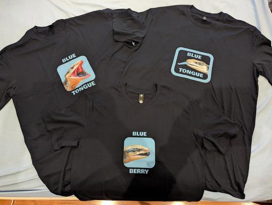 Photo of three dark blue T-shirts laid out on a bed. Each t-shirt features a different printed photo of a blue tongue skink's head.