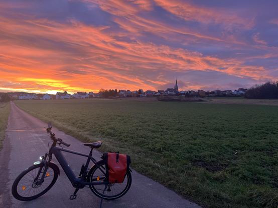 Herrliches Morgenrot (geln, orsnge, rosa, violett) über einer Dorf-Silhouette mit Kirchturm. Vorn steht ein E-Bike mit roter Satteltasche auf einem asphaltierten Feldweg. Rechts davon ein Feld.