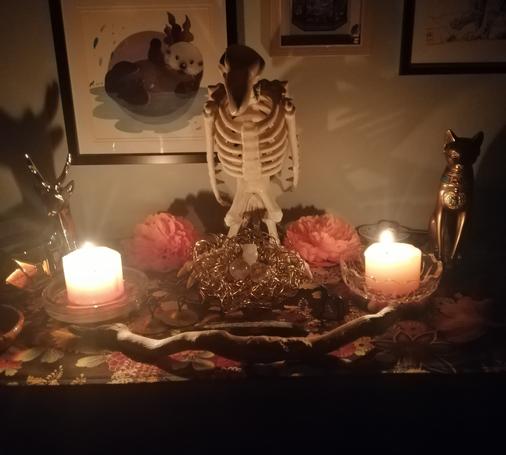 A pagan altar lit by candles. In the centre, a skeletal vulture watches over a nest of metal wire. To Her left, a silver reindeer statue. To Her right, a bronze ancient Egyptian cat statue. In front, a wand of driftwood and hints of other tools.