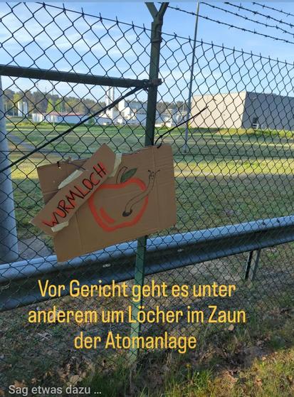 Zaun der Brennelementefabrik mit Gebäude in Hintergrund,  Zaun mit "wurmloch", Pappkarton am Zaun mit Apflel und Zaunloch drauf gemalt