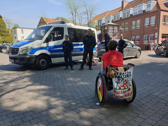 Rollstuhlfahrerin vor Polizisten, die eine Person kesseln. Banner am Rollstuhl " überall Polizei nirgendwo Gerechtigkeit " , sie trägt ein rotes T-Shirt mit atom- Totenkopf