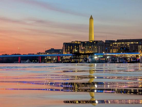 The illuminated Washington Monument at dusk with pink light on the horizon and the 395 bridge lit in blue and red all reflect in the Washington Channel and light ripples in the water