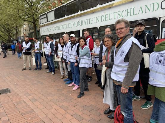 Schräg von der Seite: mindestens 14 Leute mit Hamburg-Werbefrei-Westen stehen auf der gepflasterten Fußgängerzone vor dem Omnibus für Direkte Demokratie in Deutschland.