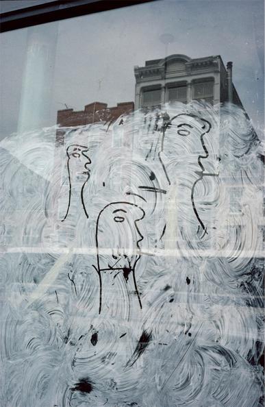 Color photograph: A window is whitewashed opaque, but there is a finger-drawing on it. Above the whitewash, buildings are reflected.