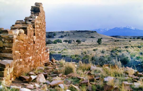 In the golden light of early evening beneath a pale gray blue sky, we are standing in front of partially excavated ancient native American pueblo ruins built of adobe bricks. The semiarid plain beyond is covered with pale gold grasses with occasional green leafy shrubs and a few trees. Numerous rocks and small boulders are visible where the grass does not grow. Small grassy ridges and hills rise in the background. Above and beyond them a range of loftier snowcapped mountains towers in the distance.