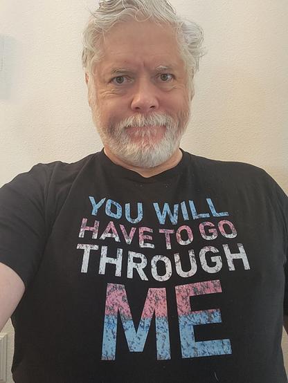A white-bearded white man in his sixties wearing a black t-shirt with the faded multicolored words "YOU WILL HAVE TO GO THROUGH ME."