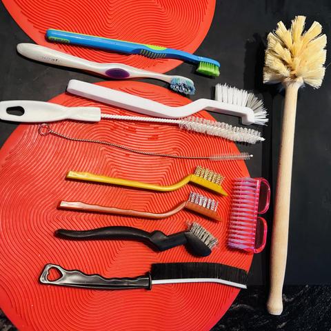 Almost a dozen cleaning tools laid out on the counter on a black silicone mat with 2 red round silicone trivet mats. The crevice tool brush is at bottom. The favorite slim white brush is 3rd down, under my standby old toothbrushes.
