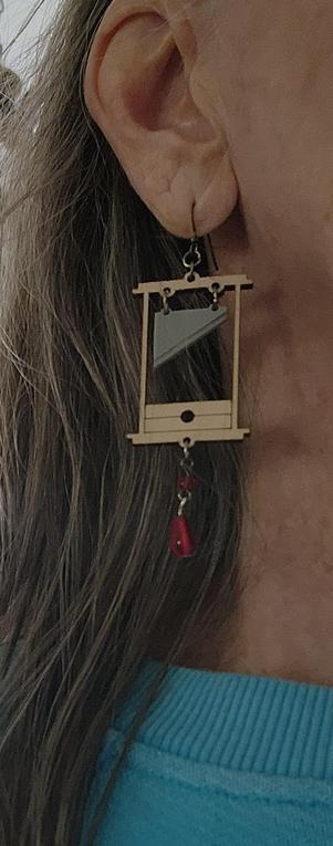 Guillotine earring. Cream colored structure with red bead droplet hanging from bottom. Blade is silver.