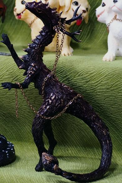 The figure of scalie creature, made from smth polymer. It portrays the bipedal reptile (?) with black scales and big horns on the head, and with golden chain wrapped around the horns and the tail base as a jewelry.