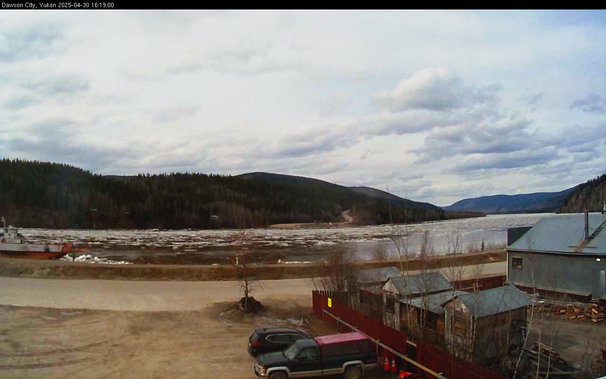 Webcam image from atop the fire station in Dawson looking across the Yukon River to the hill beyond and mostly cloudy skies above.