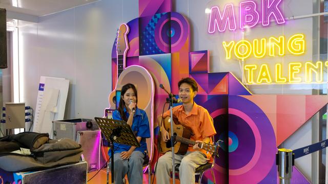 color photo of 2 street performers at a shopping center in Bangkok. A young man and a young lady sitting together with the name of the shopping center in neon light on the wall behind them where it says: "MBK Young Talents". Both performers are looking directly at the viewer. The young man dresses in a orange t-shirt and long pants, the lady wears a blue short sleeves shirt and jeans. The young man is holding a guitar singing into a standing mic while the girl holding a mic also singing.