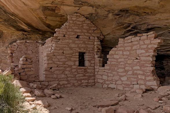 Color landscape photo showing primitive stone walls made of reddish brown sandstone. There a couple of ninety degree angles with one wall having a large opening that was possibly a door. The structure is under the roof of a sandstone alcove (shallow cave). The structure is badly eroded.