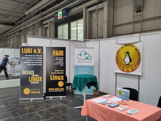 Das Bild zeigt einen Messestand von LUKi e.V. (Linux User im Kirchlichen Bereich), einer Organisation, die den Einsatz von Linux und freier Software im kirchlichen und religiösen Umfeld fördert.
Der Stand zeigt mehrere Banner mit verschiedenen Botschaften:

Ein Banner mit dem Text "Jesus würde LINUX nutzen"
Ein weiteres Banner mit "FAIR ist ein Glück" und Informationen über faire Software
Ein "LibreChurch"-Banner, das als "Modellprojekt für freiheitliche und nachhaltige" Lösungen beschrieben wird

Auf der rechten Seite ist ein rundes gelbes Logo mit dem Linux-Pinguin und dem Text "Linux User im Bereich der Kirchen - LUKi".
Am Stand befindet sich ein kleiner Tisch mit einer orangefarbenen/korallenfarbenen Tischdecke, auf dem einige Bücher oder Informationsmaterialien ausliegen. Außerdem ist ein kleines Schild zu sehen mit "Befreie Dein Android".
Die Umgebung scheint eine Messehalle oder ein Ausstellungszentrum zu sein, mit temporären Trennwänden, die den Standraum bilden.