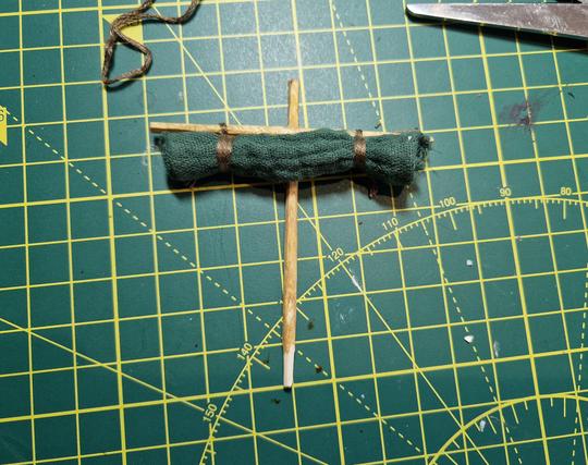 A model of a sail sat on a cutting mat. It is made from a length of cloth that has been rolled up and rigged to the horizontal yard with embroidery thread. The mast and yard are carved from long match sticks and touched up with brown ink.