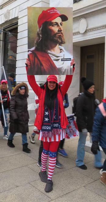 Image of a woman in US-colored clothing with various MAGA paraphernalia holding up a big picture depicting an "aryan-style" Jesus with a MAGA hat.