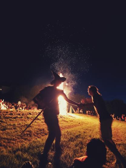 The image depicts a nighttime scene centered around a large bonfire. Sparks are visible rising from the flames against a dark sky. In the foreground, two silhouetted figures are partially visible on a grassy area. One figure on the left is wearing a hat with a pointed top and appears to be holding a stick or pole. The figure on the right is facing the bonfire. Further in the foreground, the back of a person's head and shoulders are visible. In the background, a crowd of people is gathered around the bonfire. The ground is covered in grass and is illuminated by the firelight.