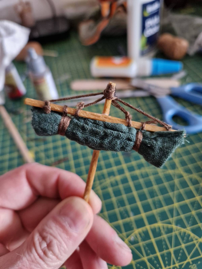 A model sail, with green cloth bundled up and tied to the yard arm. The yard itself is lashed to the top of the mast with embroidery cord to make it look like it's properly rigged. As properly as it's going to get anyway. It's being held in a white guy's hand, with the mast between thumb and forefinger. In the background is a cutting mat and crafting paraphenalia, such as superglue, scissors, and PVA glue.