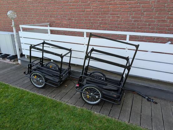 Zwei schwarze Fahrradanhänger mit je zwei quaderförmigen Aufbauten aus Aluprofilen. Die beiden Anhänger stehen auf einem Weg aus Holzplanken vor einem weißen Zaun entlang einer Rasenfläche.