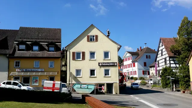 beige-creme-gelb-dreckiges Gebäude mit teils Fensterläden, Bierwerbung, Container davor, eine Straße, alles wirkt nicht mehr zeitgemäß und geschlossen