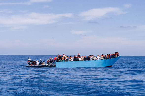 Ein blaues Holzboot voller Menschen treibt auf dem Meer. Alles sind mit Rettungswesten versorgt. Achtern liegt ein RHIB, das Menschen von dem Boot an Bord nimmt.